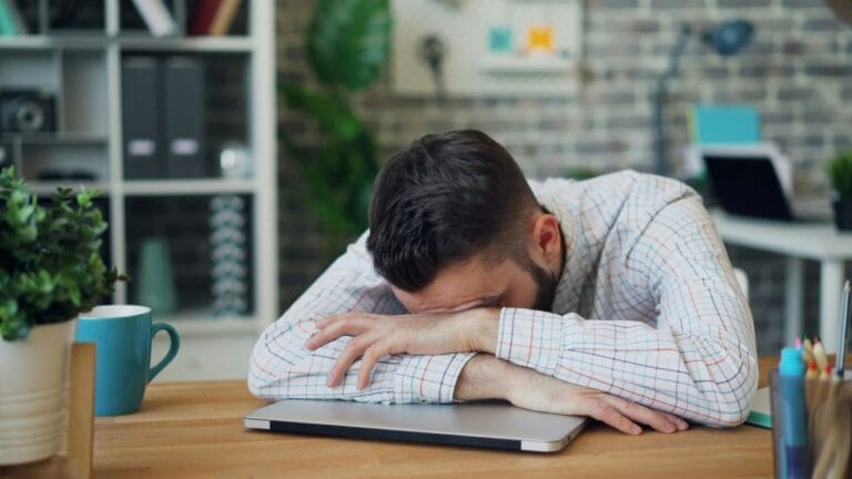 Man slaapt uitgeput op zijn bureau door slaaptekort. Hoeveel slaap per leeftijd heb je nodig?